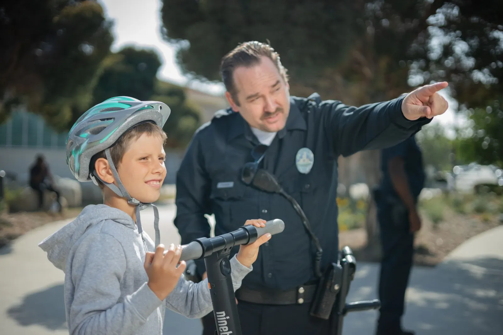 A police officer guides a young boy with a scooter outdoors, fostering community relations.