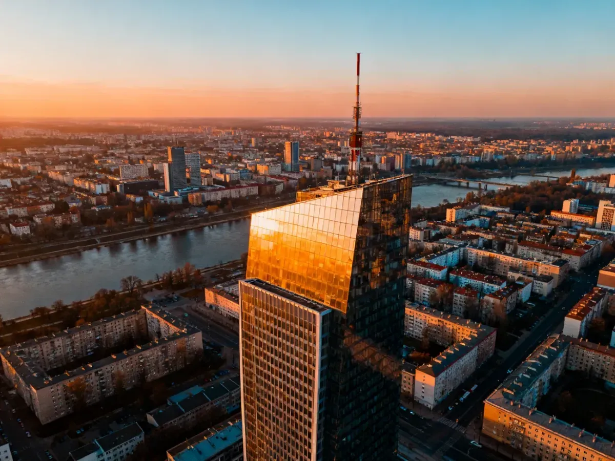 Panoramiczny widok na Warszawę o zachodzie słońca, ukazujący nowoczesne wieżowce, Wisłę oraz gęstą zabudowę miejską.