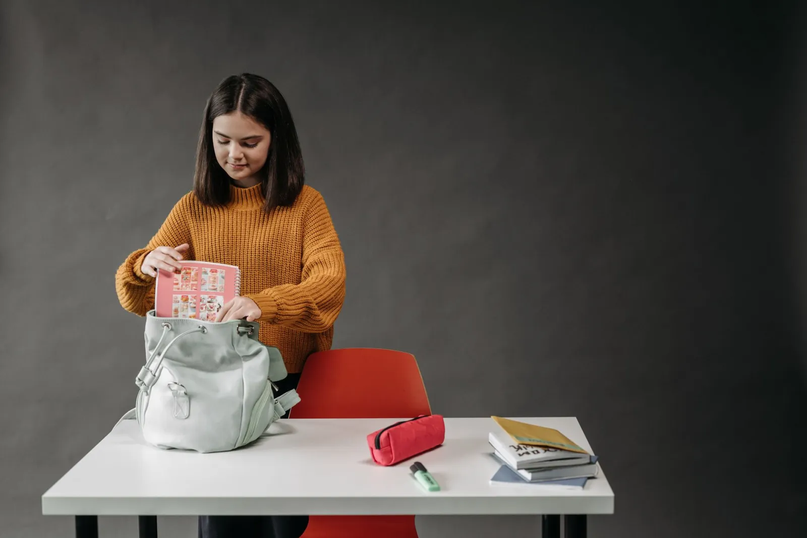 Caucasian girl organizing school supplies into her backpack at a desk.